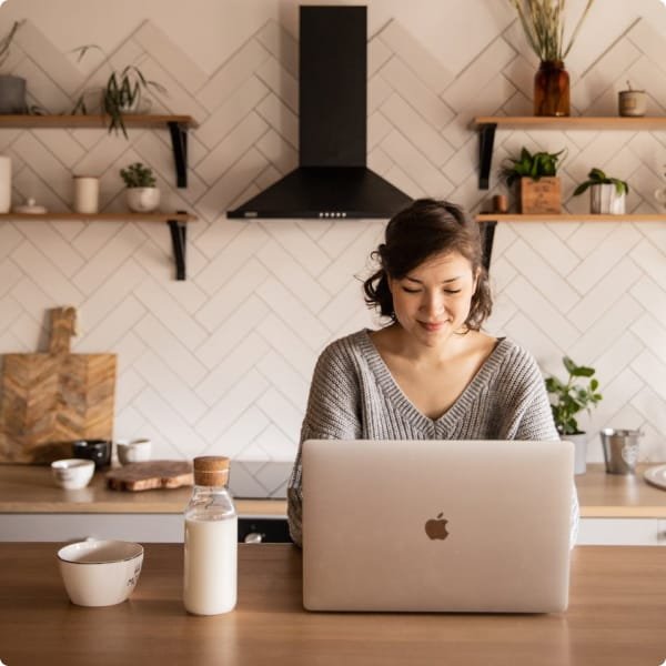 Home Smiling woman in her 40s sitting at a brown kitchen table, working on a Mac laptop. On the table are a bottle of milk and a bowl. Behind her is a white wall with kitchen utensils, plants, and pottery.